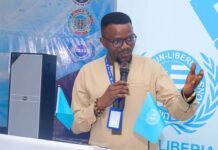 Professor Duramany-Lakkoh Strengthens IPAM’s Partnerships with LIPA, AMEU and MUN-Liberia Man in a beige shirt and glasses speaks into a microphone at a podium, with blue UN flags and a banner in the background.