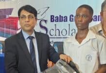 Indian Charge d’Affaires Inspires Pupils at Baba Guru Nanak Scholarship Ceremony Man in a suit hands a prize to a student in a white uniform on stage, with a banner in the background.