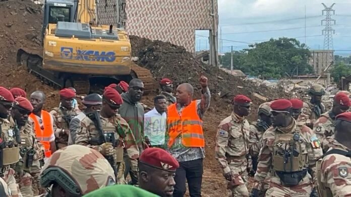 Group of soldiers in desert camouflage and red berets standing at a construction site, with a bulldozer on a dirt hill and a person in an orange safety vest gesturing to the crowd.