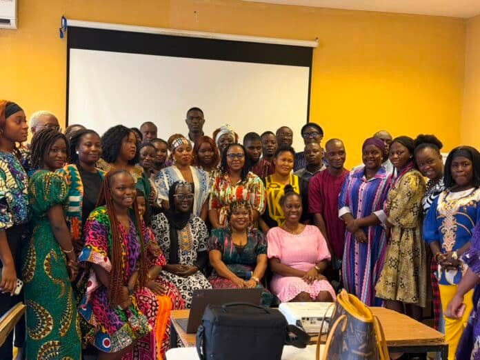 Group of men and women in colorful traditional African attire posing for a photo in a bright room with a projector screen behind them.