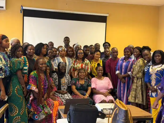 Group of men and women in colorful traditional African attire posing for a photo in a bright room with a projector screen behind them.
