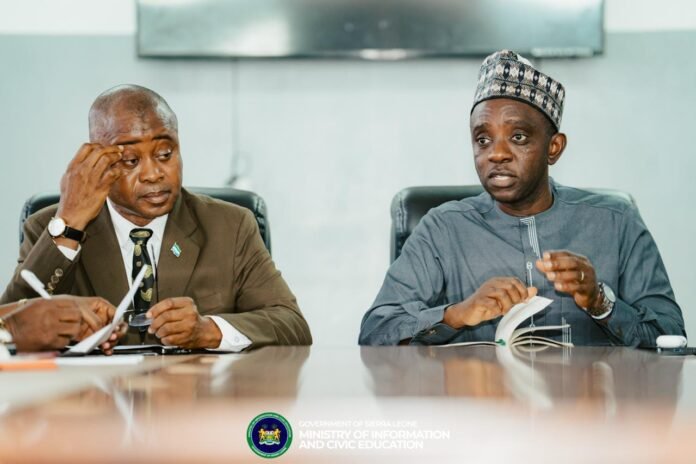 Two men sit at a conference table in a formal meeting; one in a suit rubs his temple while the other in traditional attire speaks and turns pages of a notebook. Sierra Leone government logo visible on the desk.