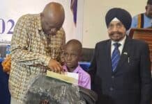 Baba Guru Nanak Scholarship 2026 Marks 5th Edition, Awards Full Scholarships to 350 Pupils A man in a patterned shirt helps a young boy hold a large, wrapped package during a formal event, with another man in a suit nearby.