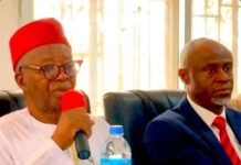 High Court Freezes APC Executive Operations, Restrains Yansaneh and Dumbuya Man wearing a red fez and glasses speaks into a red microphone at a panel, with a water bottle on the table beside him.