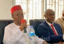 APC Rejects PR System, 50%-Plus-One Threshold as Constitutional Amendment Debate Intensifies Man in white traditional attire and red fez speaks into a red microphone at a formal meeting, with two suited men beside him.