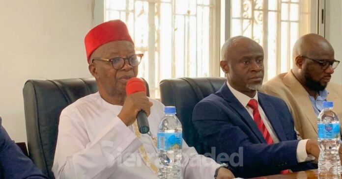 Man in white traditional attire and red fez speaks into a red microphone at a formal meeting, with two suited men beside him.
