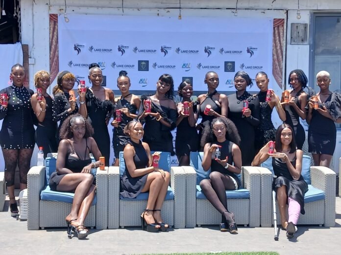 Group of women in black dresses posing with beverage cans at an outdoor event, front row seated on wicker chairs with a branded backdrop behind them.