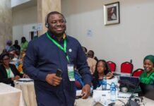 Sierra Leonean Elected VP of ECOWAS Risk Communication Network Smiling man wearing headphones and a green lanyard speaks at a conference, holding a phone, with attendees seated at tables behind him.