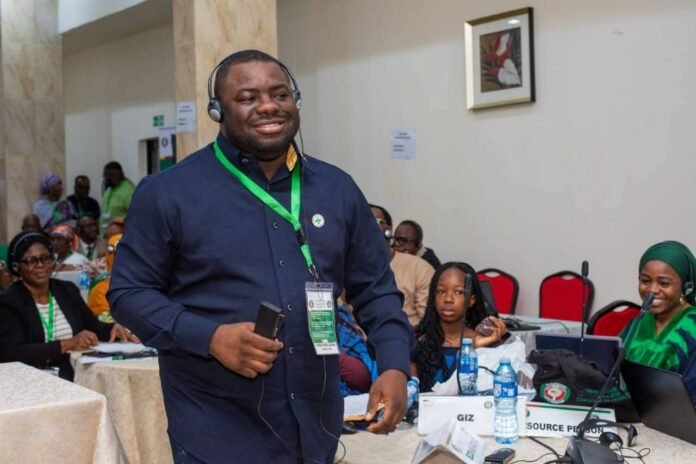 Smiling man wearing headphones and a green lanyard speaks at a conference, holding a phone, with attendees seated at tables behind him.