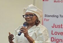 50/50 & Partners Advance Women’s Land Rights, Peace and Inclusive Governance Woman in a white traditional outfit and glasses speaks into a microphone at a podium, with an equality banner in the background.