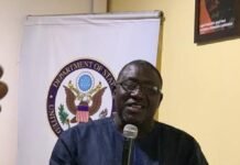 Sierra Leone Must Build DPI on Legal Identity and Institutional Collaboration — NCRA DG Man in a dark blue tunic speaks into a handheld microphone at a podium, with a Department of State banner behind him.