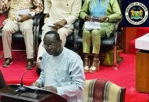 Finance Minister Says Sierra Leone Needs New Port to Unlock Economic Growth Man in a light blue traditional outfit speaks at a podium with a microphone, reading from papers; seated officials and the Sierra Leone Ministry of Finance seal visible in the background.