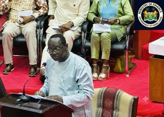 Man in a light blue traditional outfit speaks at a podium with a microphone, reading from papers; seated officials and the Sierra Leone Ministry of Finance seal visible in the background.