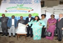 WFP Procures Over One Million Kilograms of Local Food for Sierra Leone’s School Feeding Programme Group of people at a donation ceremony standing in front of stacked bags with a banner that says HOME-GROWN school Feeding, handing a large bag of supplies to a recipient man seated center.