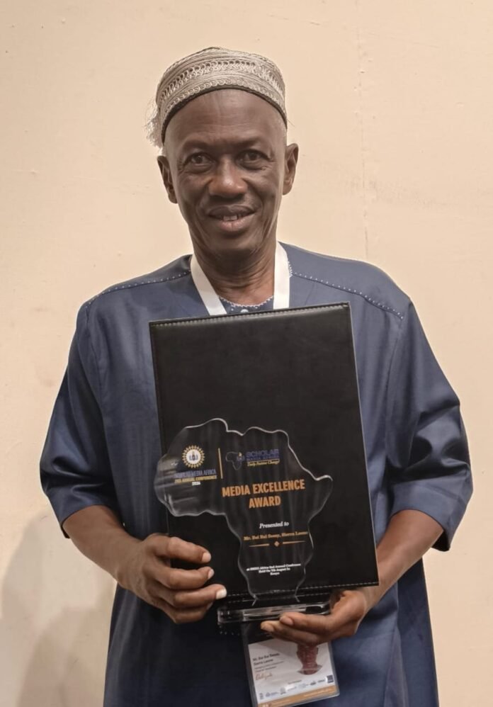 Man in a traditional cap and blue tunic holds a black award plaque titled 'MEDIA EXCELLENCE AWARD' and smiles at the camera.