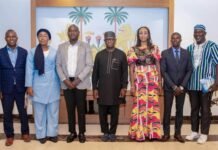 ICPNC Presents 2025 Annual Report and Five-Year Strategic Plan to President Bio Seven people in business and traditional attire posing for a group photo indoors, in front of a mural with palm trees.