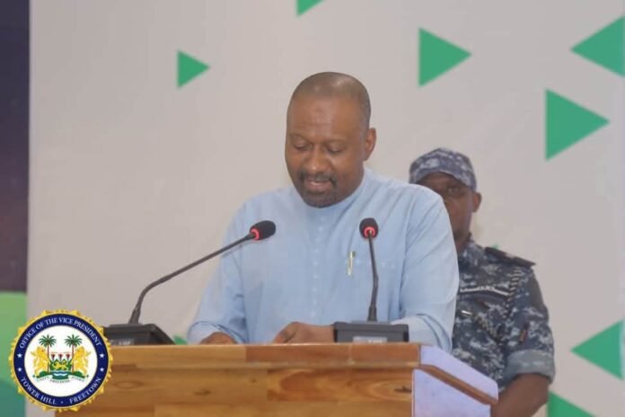 Man in a light-blue traditional shirt speaks at a wooden podium with two microphones, the VP seal visible on the front.