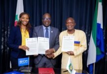ECOWAS, IOM and Partners Launch $35,000 Grant to Support Trafficking Survivors Three professionals pose with signed documents during a formal agreement ceremony, flanked by flags and a table with IOM branding at center.
