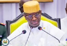 President Bio Unveils Accelerated Delivery Agenda at Opening of Fourth Session of Sixth Parliament High-ranking official in white traditional attire and a yellow cap speaks into two microphones at a formal event, with an official seal nearby.