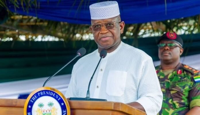 Man in white traditional attire and cap speaks at a podium with a presidential seal; a uniformed officer stands in the background.