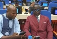 APC Mohamed Bangura, Alfred Thompson Break Ranks as Opposition Walks Out of Parliament Two men in formal attire seated at a conference hall, looking at their smartphones with blue chairs and laptops in the background.