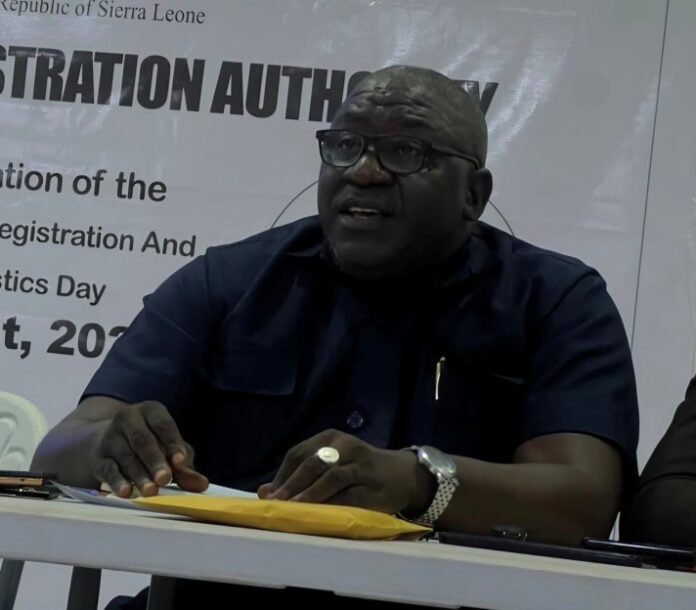 A bald man with glasses in a navy shirt sits at a table handling papers at a registration event backdrop.