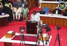 Parliament Approves 2026 Supplementary Budget to Protect Economy Amid Global Oil Shock Speaker at a wooden podium delivering a speech in a government chamber, with journalists and an audience seated behind and camera crews recording. Sierra Leone Ministry of Finance seal visible in the corner.