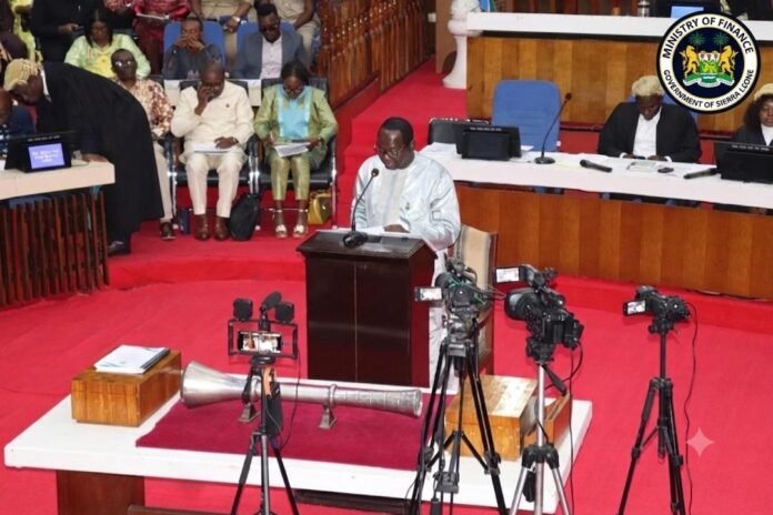 Speaker at a wooden podium delivering a speech in a government chamber, with journalists and an audience seated behind and camera crews recording. Sierra Leone Ministry of Finance seal visible in the corner.