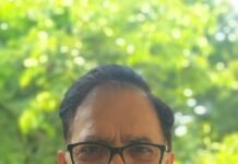 Citizen-Centric Diplomacy of India Head-and-shoulders portrait of a man with black glasses, short dark hair, and a dark textured vest, smiling outdoors among green foliage.