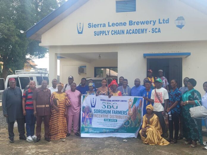 Group of men and women posing outside a cream building labeled Sierra Leone Brewery Ltd - Supply Chain Academy, holding a banner for a Sorghum Farmers Incentive Distribution event (2026).