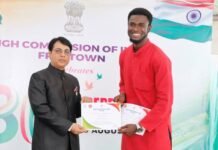 Sallu Kamuskay Receives Two Certificates and Prizes from Indian High Commission in Sierra Leone Official in a black suit presents a certificate to a smiling man in a red kurta at an award ceremony, with a banner and Indian flag in the background.