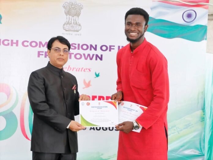 Official in a black suit presents a certificate to a smiling man in a red kurta at an award ceremony, with a banner and Indian flag in the background.