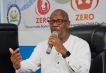 Health Minister Demands Accountability Over Preventable Maternal and Child Deaths Man in a white shirt and glasses speaks into a handheld microphone at a panel, with a banner about Zero preventable deaths in the background.