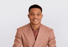 Heirs Life Assurance Appoints Pastor Jerry Eze as Independent Non-Executive Director Man in a peach-colored double-breasted suit jacket leaning on a white table and smiling at the camera.