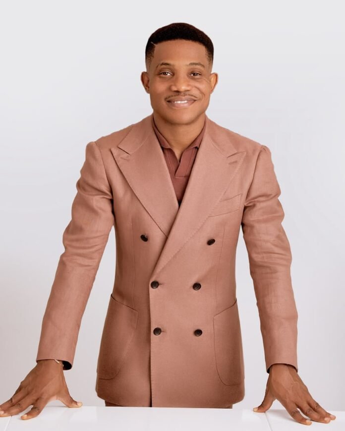 Man in a peach-colored double-breasted suit jacket leaning on a white table and smiling at the camera.