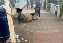 Government Urged to Take Immediate Action Against Growing Street Begging in Freetown People sit on a sidewalk beside a littered street; a woman with a child, others standing in the background, with a blue trash bin on the left and a white fence on the right.