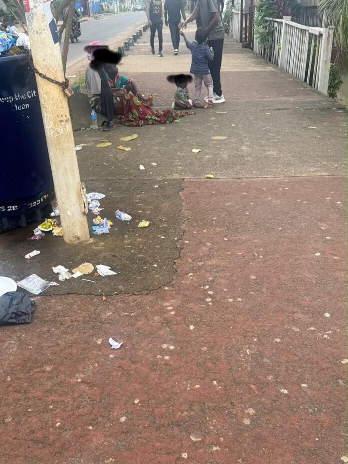 People sit on a sidewalk beside a littered street; a woman with a child, others standing in the background, with a blue trash bin on the left and a white fence on the right.