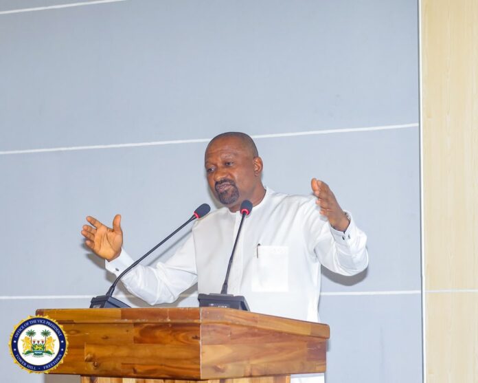 Man in a white shirt speaks at a wooden lectern with two microphones, gesturing with his hands