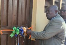SLURC Hands Over Climate Resilient Housing Upgrades in Moyiba Man in a gray suit ties colorful bow ribbons to a wooden door, likely for a ribbon-cutting or decoration event.