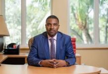 Sierra Leone’s Professor Charles C. Jalloh Secures Broad International Backing for ICJ Election Professional man in a blue suit sits at a wooden desk in an office, hands folded, with a window and greenery in the background.