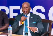 JM Mining’s Licence Offer Lapsed Over Unpaid US$1.1 Million Fees, Government Says Man in a teal suit speaks into a handheld microphone at a panel, seated behind a table with a water bottle and backdrop banner behind him.