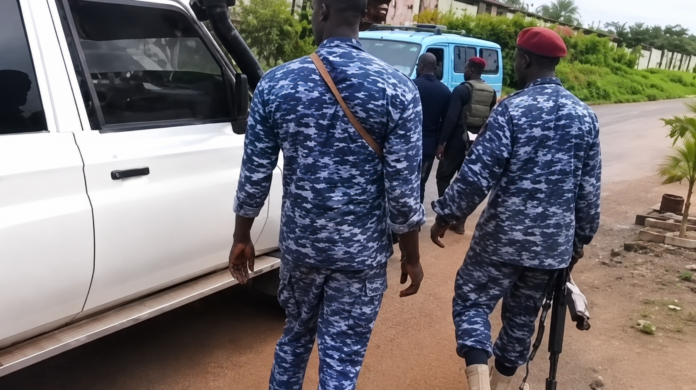 Group of soldiers in blue camouflage uniforms walking along a dusty road beside a white vehicle; one man wears a red beret and carries a rifle nearby.