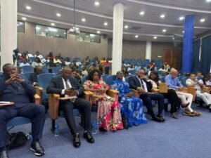 Audience seated in a large conference hall listening to a presentation, some taking notes, water bottles on floor nearby