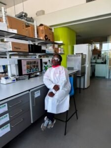 Lab technician in a white coat and pink scarf sits on a chair by a workbench with shelves and a computer monitor behind him.