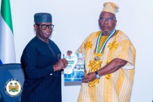 Two men in formal dress exchange a copy of a booklet titled 'Annual Report 2025' as a flag stands nearby.