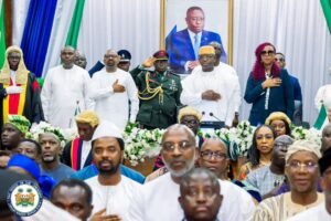 Officials and guards stand with hands over their hearts at a formal ceremony, a portrait of the president in the background.