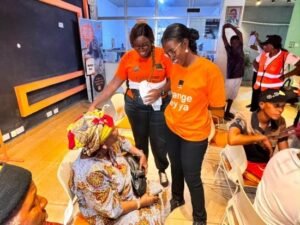 Two volunteers in orange shirts assist an elderly woman seated at a community event indoors, while others watch nearby.