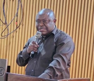 | The Calabash Newspaper Man in a dark shirt speaks into a handheld microphone at a wooden-paneled backdrop, standing behind a lectern