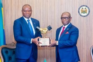 | The Calabash Newspaper Two men in navy suits shake hands and present a gold fist-shaped trophy in a formal award ceremony, with a government seal on the wall and a flag nearby.