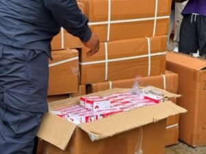 Worker in a dark blue uniform packs and stacks large taped cardboard boxes, with an open box in the foreground containing smaller red-and-white packages.